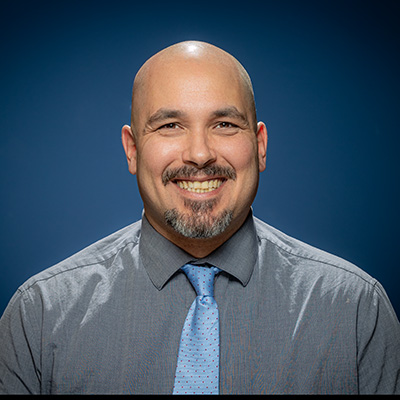 Kris LeBlanc smiling, wearing a grey shirt and blue tie
